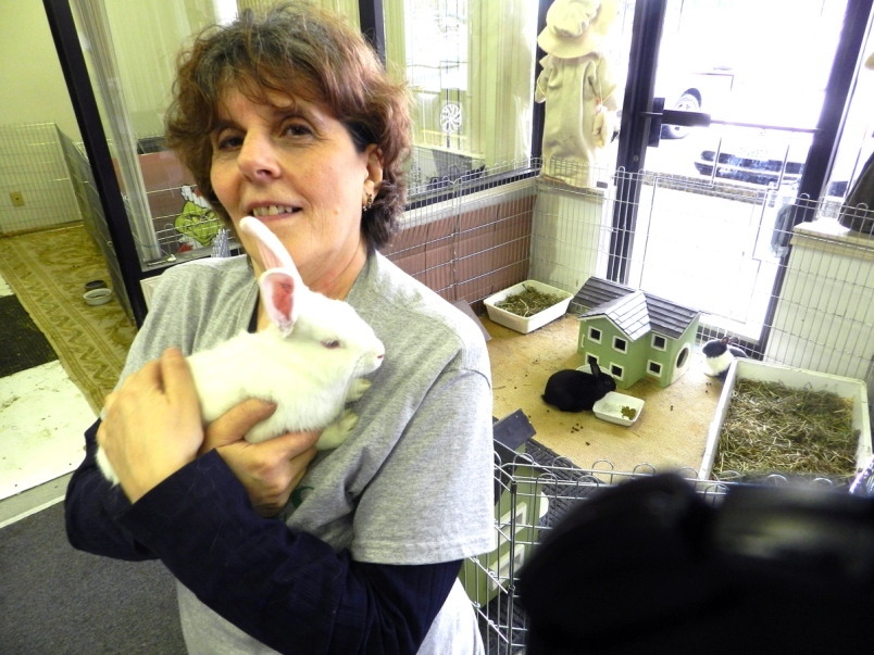 A white woman with short brown hair stands in front of a fenced off area for bunnies, holding a fluffy white bunny in her arms